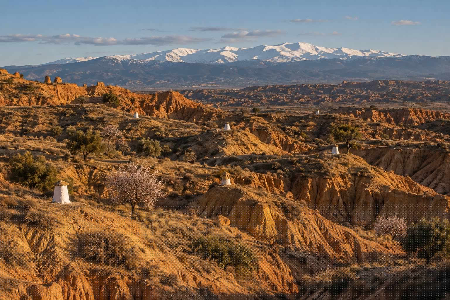 Colinas de arcilla ocre del altiplano de Guadix con chimeneas blancas de casas cueva asomando del terreno y Sierra Nevada nevada al fondo (imagen ilustrativa)