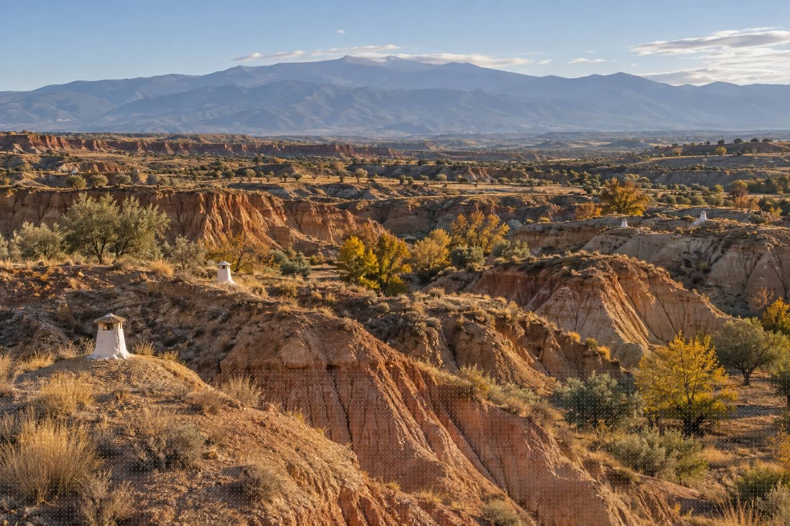 Cárcavas de arcilla ocre del Altiplano de Guadix en otoño, con chimeneas blancas de casas cueva asomando del terreno y Sierra Nevada al fondo (imagen ilustrativa)
