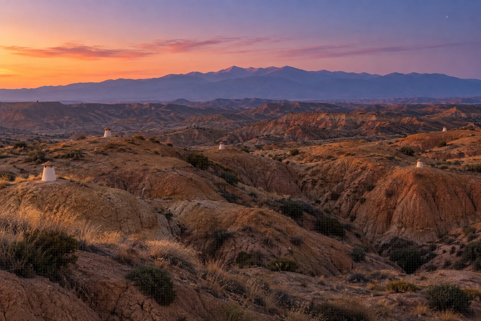 Cerros de arcilla ocre del altiplano de Guadix al atardecer, con chimeneas blancas de casas cueva asomando del terreno y Sierra Nevada al fondo (imagen ilustrativa)