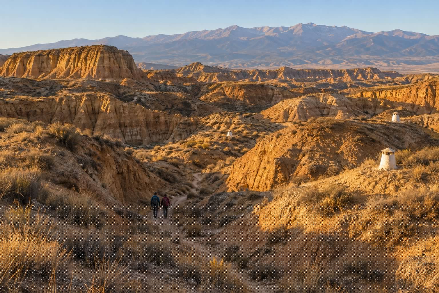 Cárcavas de arcilla ocre del Geoparque de Granada al amanecer, con chimeneas blancas de casas cueva asomando del terreno, dos senderistas de espaldas en un camino y Sierra Nevada al fondo (imagen ilustrativa)