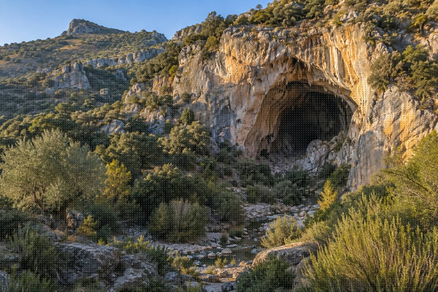 Boca de una cueva natural en un cantil de roca caliza de una sierra andaluza, orientada al sur, rodeada de matorral mediterráneo y con un cauce en el valle