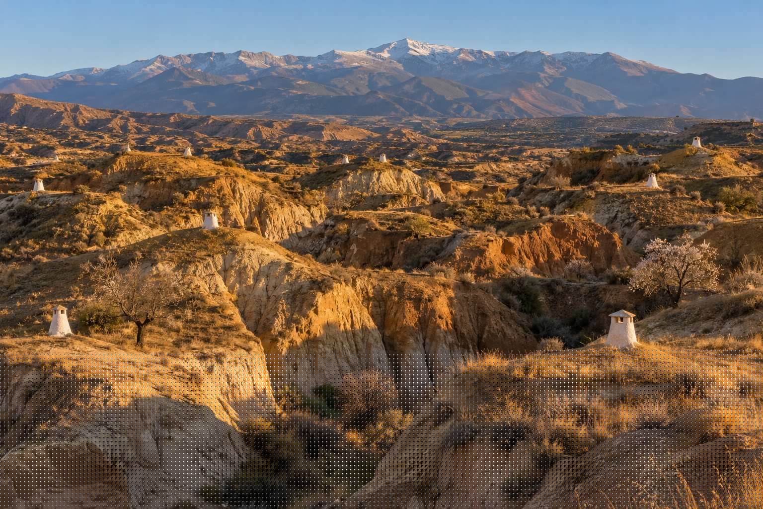 Cerros de arcilla ocre del altiplano de Guadix con chimeneas blancas de casas cueva asomando del terreno y Sierra Nevada al fondo, con luz de atardecer de otoño (imagen ilustrativa)
