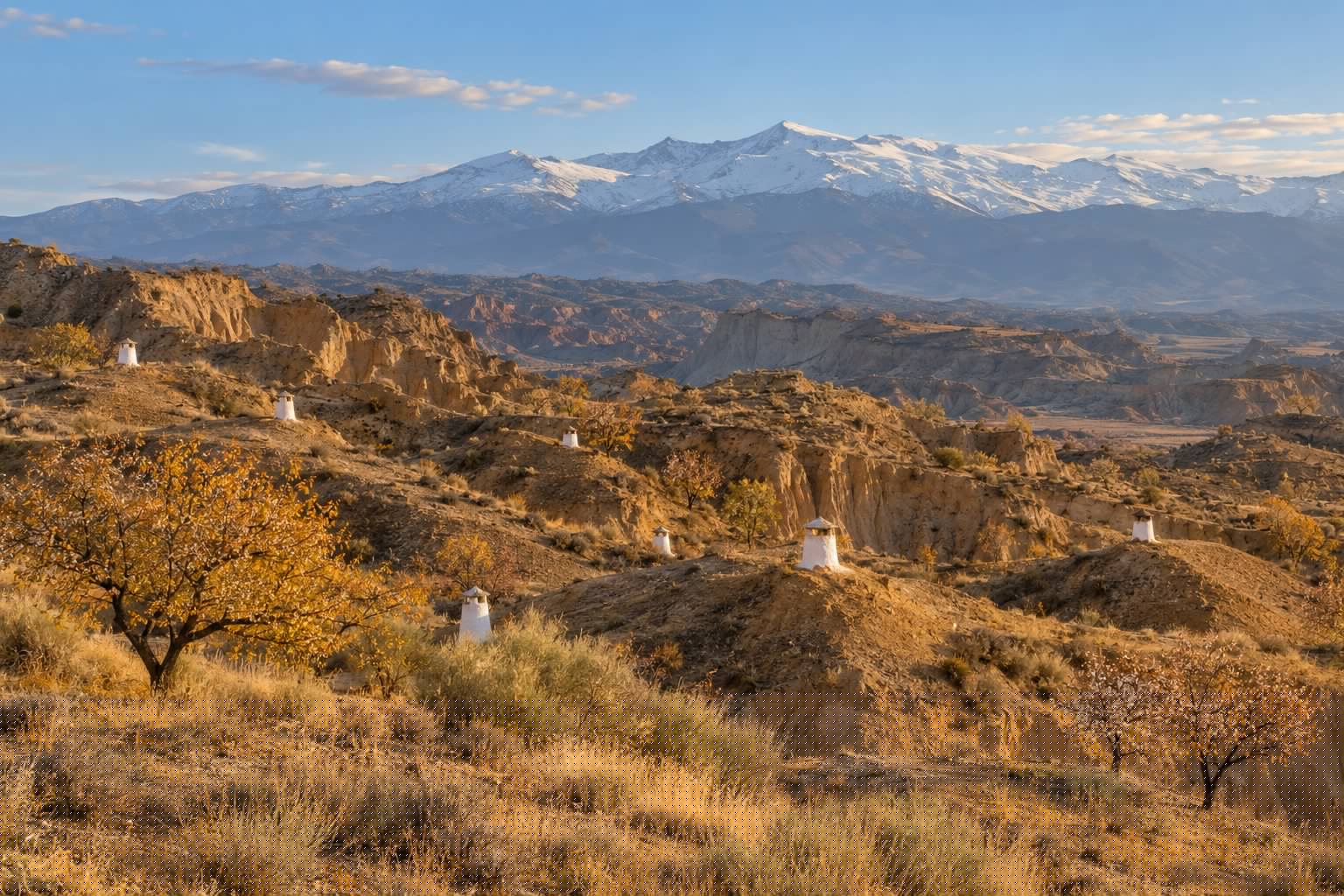 Lomas de arcilla ocre del altiplano de Guadix con chimeneas blancas de casas cueva asomando del terreno y Sierra Nevada al fondo, con luz dorada de otoño (imagen ilustrativa)